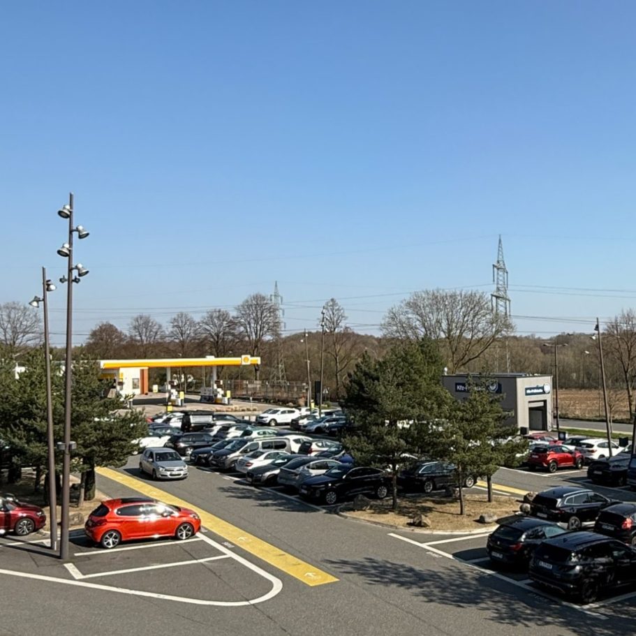 Parkplatz des Ruhrparks mit vielen Autos und einem Tankstellengebäude (Shell) und einer KFZ-Prüfstelle im Hintergrund.